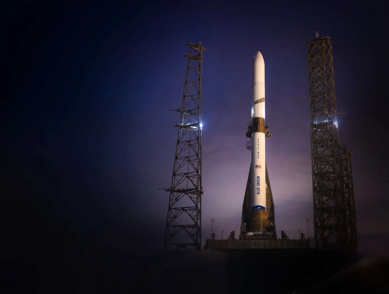 A Blue Origin New Glenn rocket stands on a launch pad at dusk, flanked by illuminated towers against a deep blue sky, conveying anticipation and technological progress.