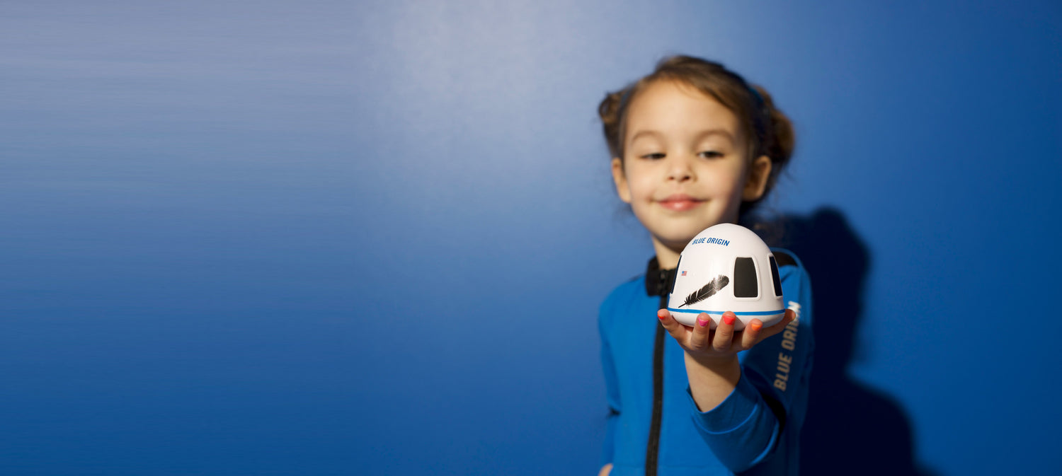 A child in a kids Blue Origin Space suit outfit holding a small model of a Blue Origin New Shepard Capsule against a blue background, illustrating interest in space exploration and STEM education.